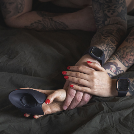 A couple lying in a bed with dark sheets hold hands. She also has the Pulse stroker in one of her hands.