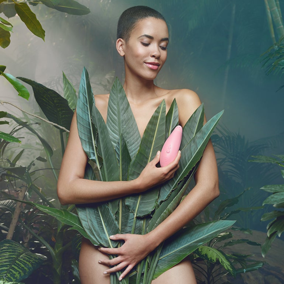 A woman of colour with short hair poses nude in a forest with leaves used to conceal her intimate areas. In her hand she holds the Womanizer Premium Eco, a light pink curved pleasure device with two visible buttons.
